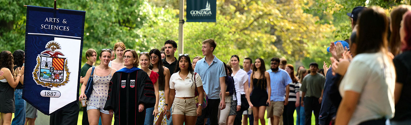 A large group of students walking behind an Arts & Sciences flag.