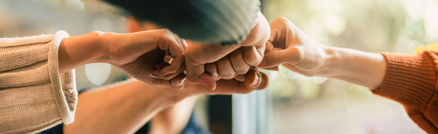 A close-up, horizontal shot of four people's fists gently bumping together in the center of the frame, signifying teamwork and collaboration.