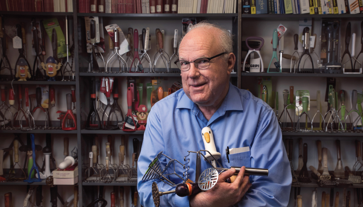 Older man with blue shirt holding potato mashers standing in front of a bookshelf lined with hundreds of potato mashers