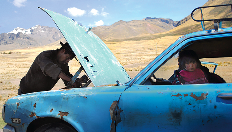 A man works under the hood of an old car while a child sits in the driver's seat, watching. Somewhere in South America.