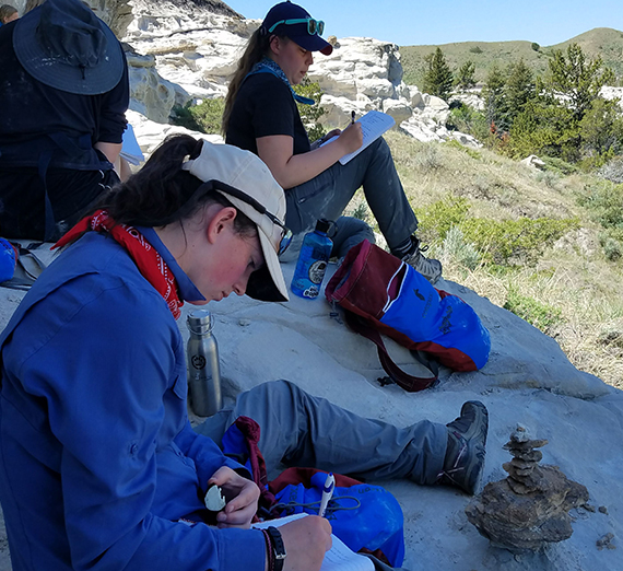 Students sit in the shade of a cliff while journaling outdoors. 