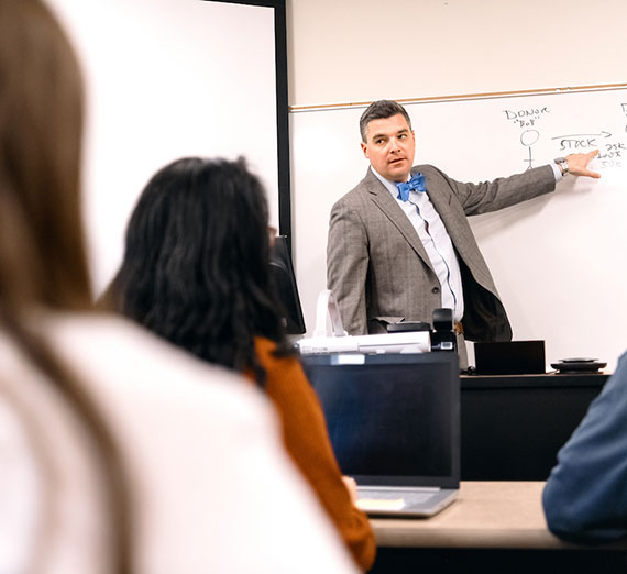 A professor stands at the whiteboard in a graduate-level business course as students listen
