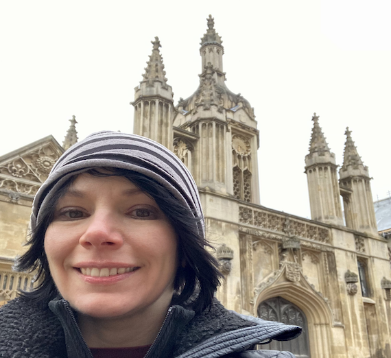 Melody Alsaker wearing hat and jacket, taking a selfie in front of older building at Cambridge University.