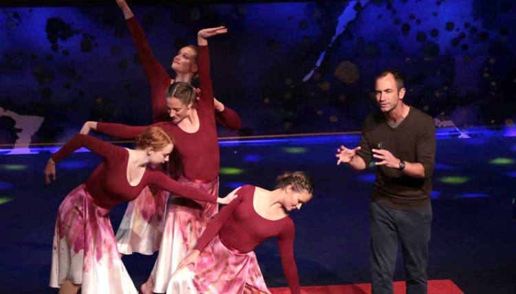 Four dancers in red long sleeved shirts and pink/white skirts enact dance that resembles the movements of animals. Professor Brook Swanson speaks to a crowd in front of them.