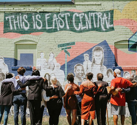 A group of people with their arms around one another standing in front of a mural that displays community members and "This is East Central". 