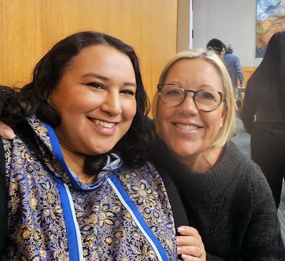 Two women smiling closely together indoors, one wearing a stethoscope, suggesting a friendly moment between healthcare colleagues.