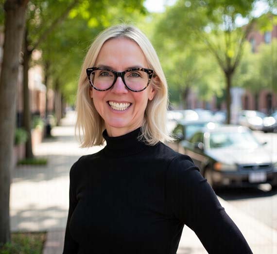 Alexis, with blonde hair, a black turtleneck and brown glasses on a sunny street.