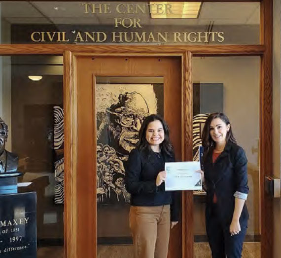Two women stand smiling in front of a door labeled "THE CENTER FOR CIVIL AND HUMAN RIGHTS," with one holding a certificate.
