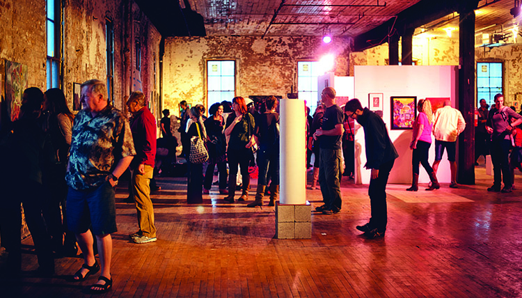 A group of spectators look at a variety of art exhibits in a brick room with wood floors. 