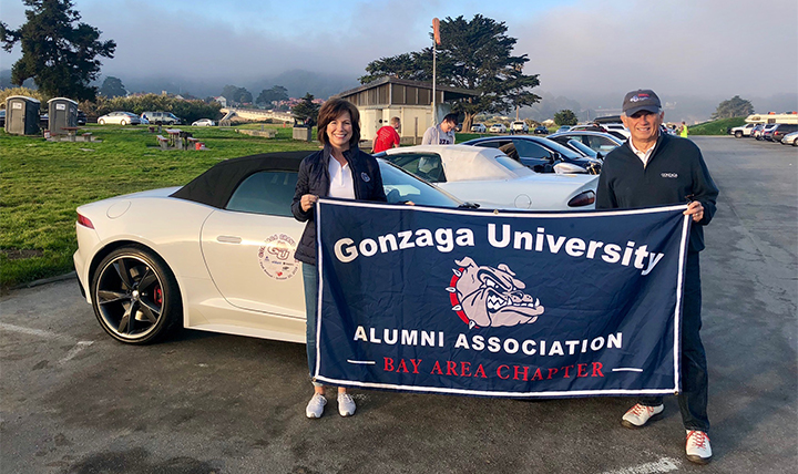 Two members of the Gonzaga Grand Prix hold a Gonzaga alumni flag in front of their car.