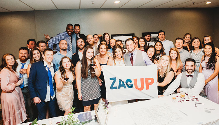 A group of wedding guests indoors, smiling and holding a 'Zag Up' flag.