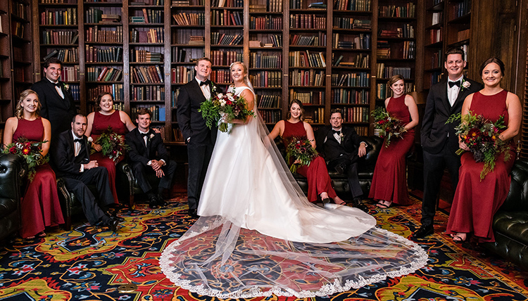 A bride and groom standing in a library, surrounded by books and in the company of bridesmaids - in red dresses - and groomsmen - in black suits.
