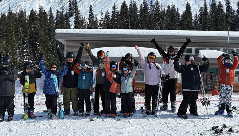 A group of Indigenous youth skiers pose in front of treed, snowy mountain