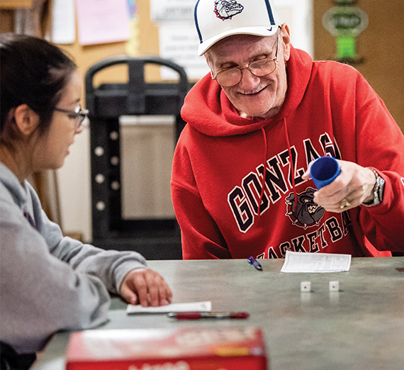 An elderly man wearing a Gonzaga hoodie plays a dice game with a student. 