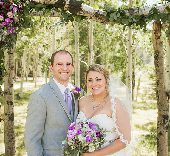 A man in a grey suit and purple tie stands under an outdoor wedding arch  next to his bride, who's wearing a wedding dress and holding a  bouquet.
