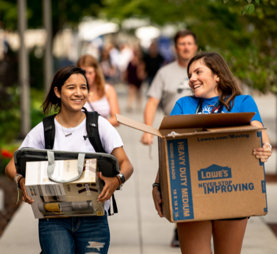 Two girls walking along a concrete sidewalk carrying packing boxes. The girl on the left side of the photo is wearing a white tee shirt with jeans and carrying a box and a black backpack. The girl on the right is wearing a blue tee shirt and black shorts. Both girls are smiling at each other. 