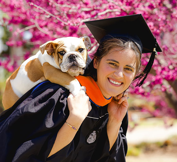 Woman in graduation cap and gown leaning with a bulldog puppy on her back