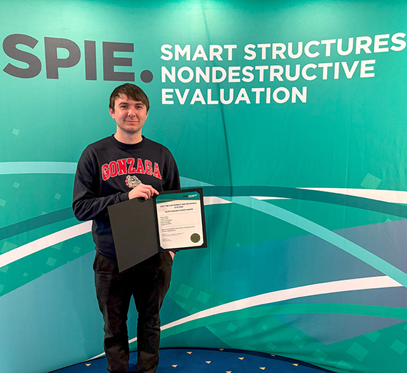 Male wearing Gonzaga sweatshirt holding an certificate award in front of a conference backdrop