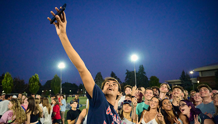 A man holds up a cell phone to take a selfie in front of a group of students outside under a dark sky