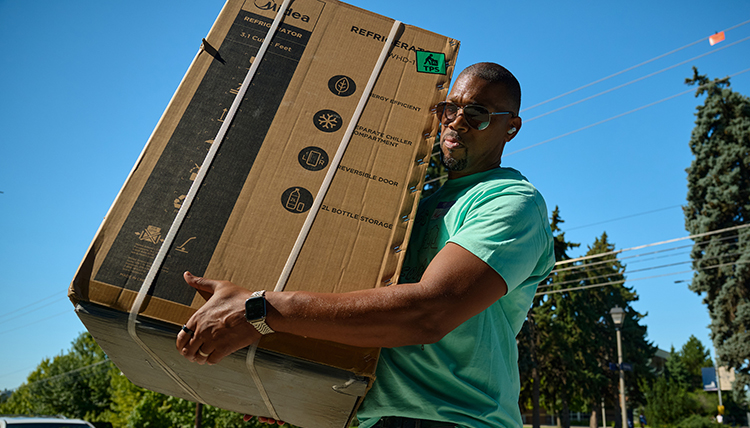 A man wearing a teal t-shirt carries a large box