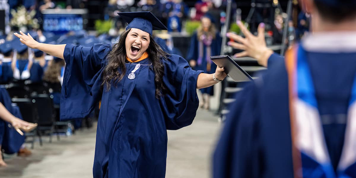 A smiling woman with her arms wide open, holding a degree in one hand, wearing a graduation cap and gown