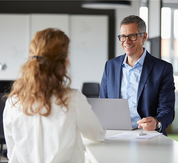 A man in glasses and a sport jacket with a computer in front of him, talking to a health care professional