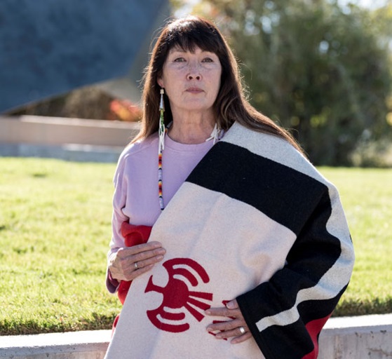 Charlene Teters standing in a field wrapped in a blanket. 