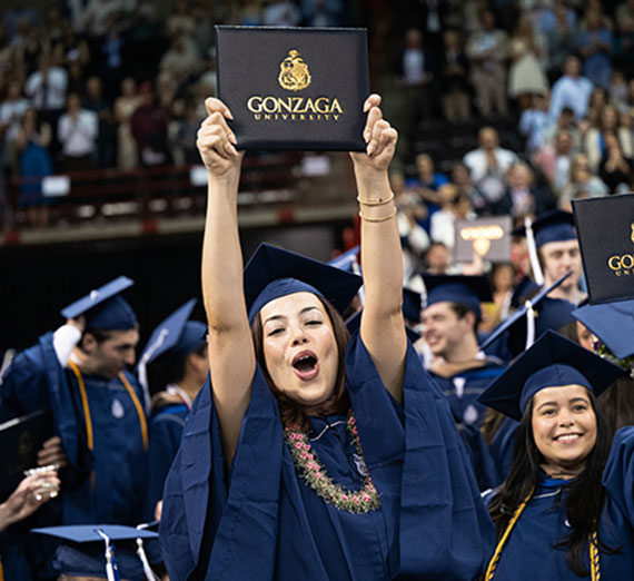 A female student in graduation cap and gown holds her degree over her head. 