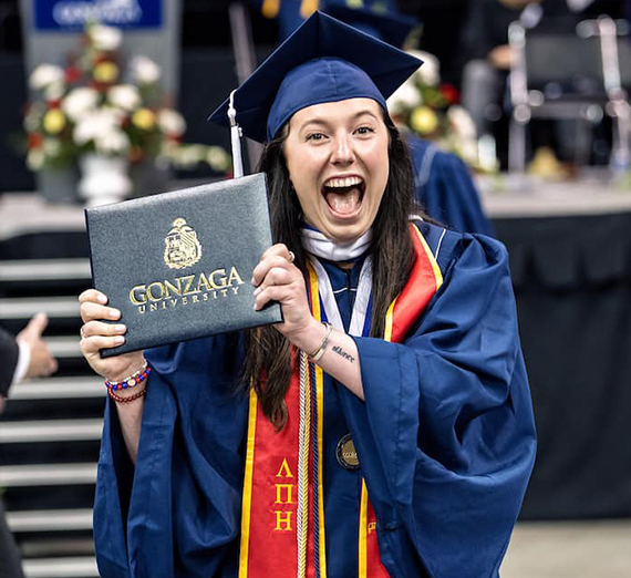 A woman in graduation cap and gown holds up her degree and smiles