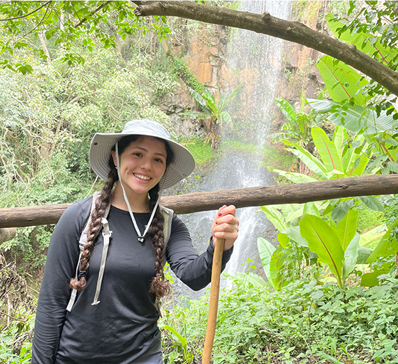 Ana Reyes standing in front of a waterfall, holding a walking stick in one hand, and wearing a sun hat
