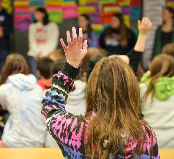 Elementary student from the back in a classroom with her hand raised.