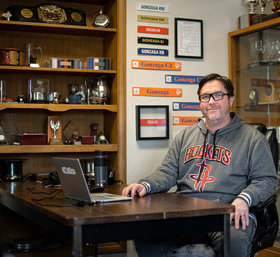 GU debate coach Glen Frappier sitting at a desk with a bookshelf of trophies behind him