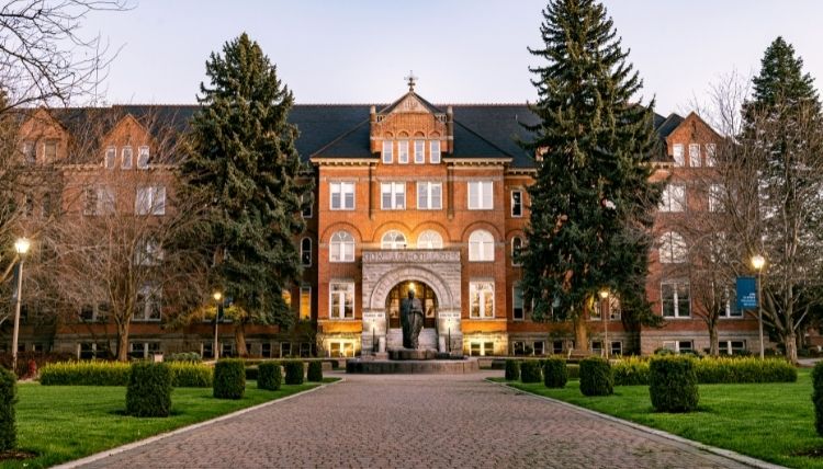 A shot of the front of College Hall, including the statue of St. Ignatius in front of the main entrance. Trees surround the building. The sun is reflecting in the windows.