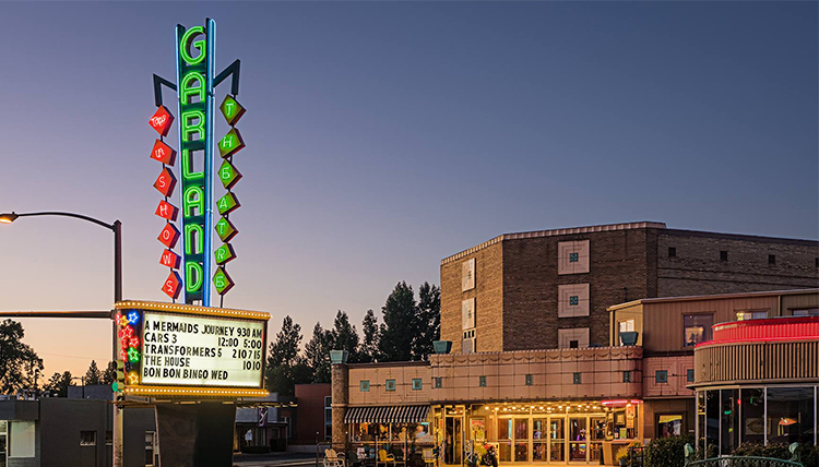 The neon sign of the Garland Theater is lit up at night and lists the movies being shown. 