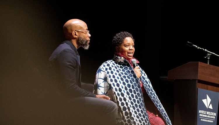 Woman with microphone talking onstage, next to man who is listening as they lead a discussion together