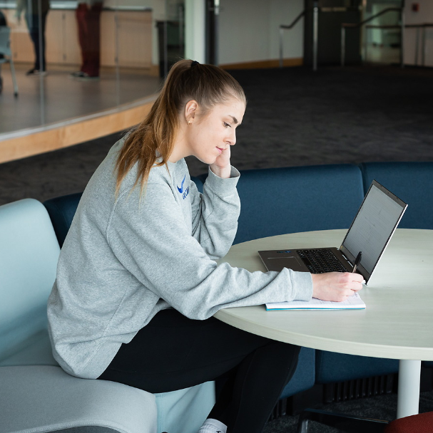 A female student studying in the Bollier Center. The student is sitting on a gray seat leaned over a laptop and a notebook. She is writing on the notebook with a black pen. She is wearing a grey long-sleeve sweatshirt with a blue logo. She is also wearing black leggings with white socks. Her hair is in a ponytail and she looks calm and is smiling slightly.