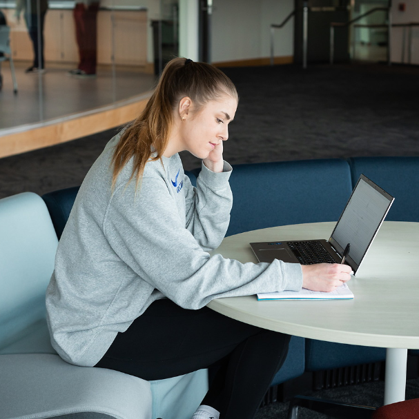 A female student studying in the Bollier Center. The student is sitting on a gray seat leaned over a laptop and a notebook. She is writing on the notebook with a black pen. She is wearing a grey long-sleeve sweatshirt with a blue logo. She is also wearing black leggings with white socks. Her hair is in a ponytail and she looks calm and is smiling slightly. 