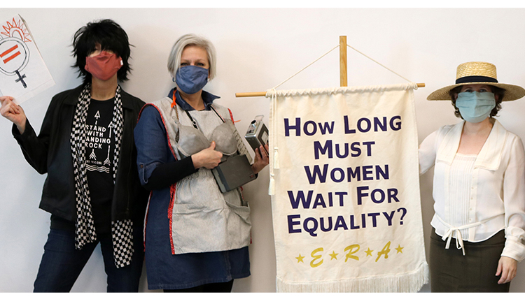 three women in costumes and a banner that reads How Long Must Women Wait for Equality