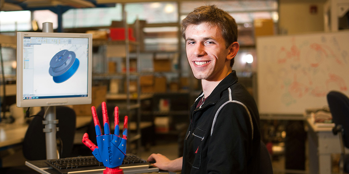 Gonzaga alum Christopher Birmingham in front of a computer screen, holding a robot hand he created