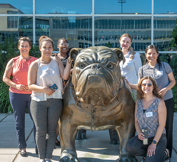 6 women posing with the bronze Spike statue in front of the athletics center