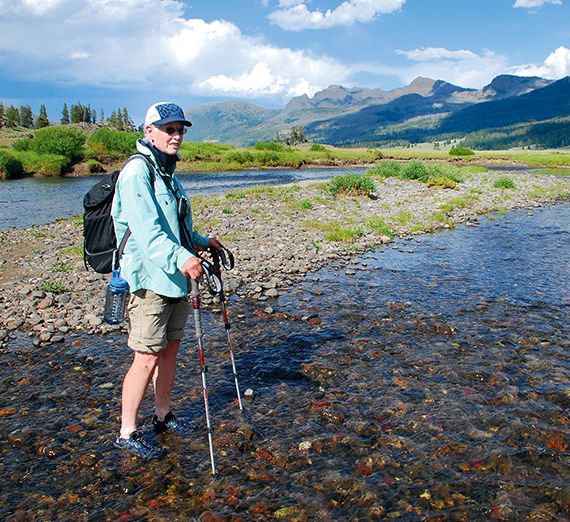 A woman wearing shorts, a sweater and a hat, holding hiking poles, standing in a creek with mountains in the background