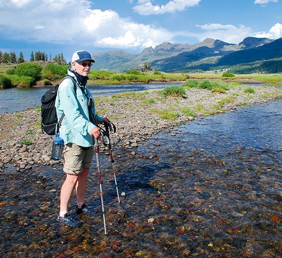 A woman wearing shorts, a sweater and a hat, holding hiking poles, standing in a creek with mountains in the background 