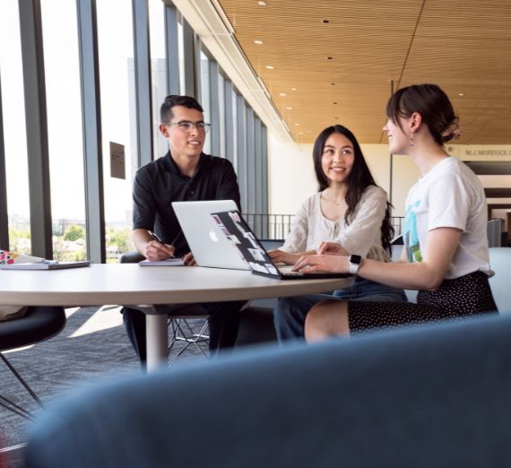 Three students sit at a table with laptops in the Bollier Center for Integrated Science and Engineering.  They are smiling at one another.