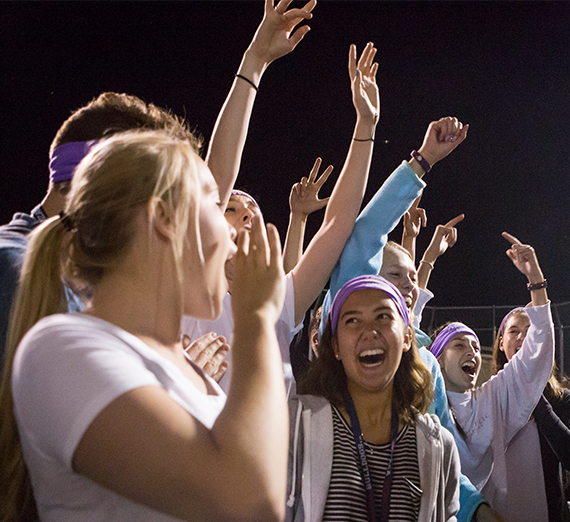 A group of students stands outside at night, cheering with arms raised.