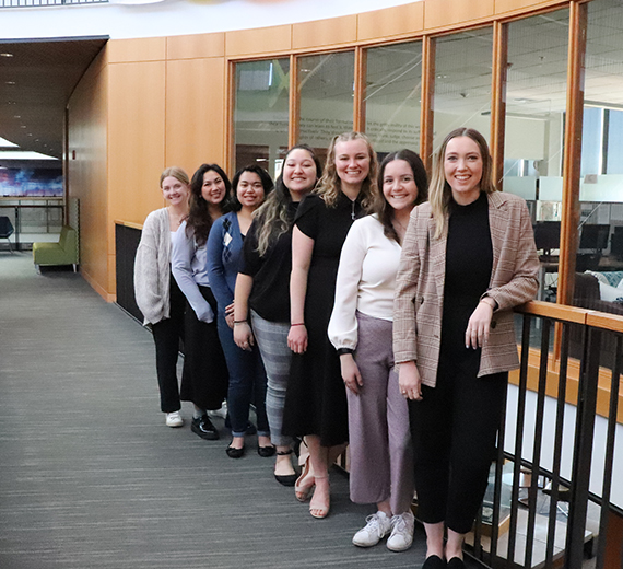 Seven Opportunity Northeast fellows pose on the walkway in the Hemmingson rotunda. They are dressed in business casual. 