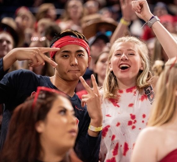 Students posing at a basketball game. The student on the left has peace signs up and the student on the right has an arm up, cheering.