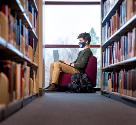 A student studying in a red chair in Foley Library. The photo is taken from an aisle of books. 