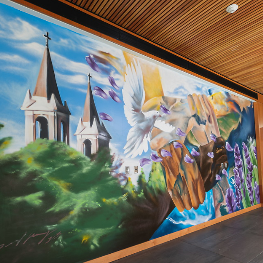 Photo of the Unity Multicultural Education Center mural that was painted in Spring 2023 in Gonzaga University's Hemmingson Center. The mural pictured has on the left a pictures of church spires above green foliage. There are two spires which are white and beige with crosses on top. In the middle of the mural is a white dove in the foreground. Then on the right  half of the mural are four interlocking arms holding each other. Each arm is a different skin tone. Then, beneath and to the rightside of these arms is a river flowing and lilac flowers. Lilac petals are blowing across the scene in the mural.