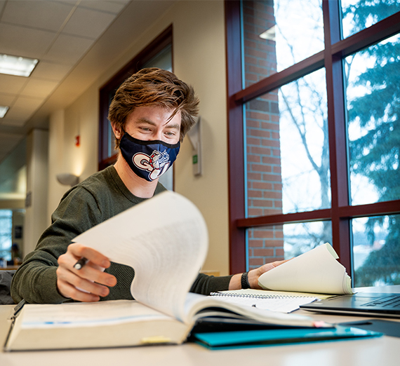 A student looks through a textbook and types on their computer while wearing a mask.