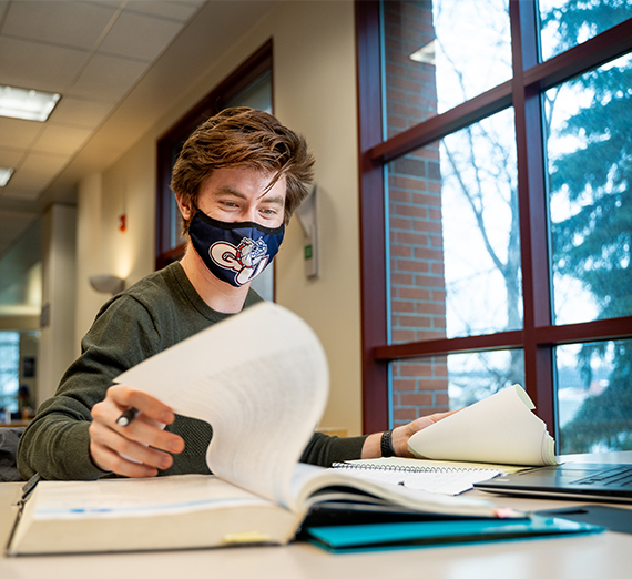 A student looks through a textbook and types on their computer while wearing a mask. 